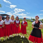 Członkinie jednego z Kół Gospodyń Wiejskich w białych koszulach i czerwonych sukienkach pozujące do zdjęcia na zielonej trawie na tle gór i błękitnego nieba.
