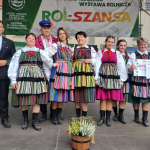 Grupa osób ubrana na ludowo i mężczyzna w garniturze pozuje wspólnie na scenie do pamiątkowego zdjęcia