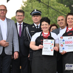 Dwie uśmiechnięte kobiety w białych bluzkach, czarnych fartuchach i czerwonych koralach stoją na podium; jedna trzyma dyplom za II miejsce, druga czek na 1500 zł, w tle  patronami i organizatorzy wydarzenia