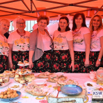 Siedem uśmiechniętych kobiet w średnim wieku z Koła Gospodyń Wiejskich Zwierzyniec Pierwszy pozuje do zdjęcia za stołem z regionalnymi potrawami; na stole plakietka „Bitwa Regionów – pierwsze miejsce”, kobiety w kwiecistych, ludowych spódnicach i bluzkach z imionami 