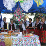 Siedem seniorek z Koła Gospodyń Wiejskich w Lgocie Murowanej, ubranych w tradycyjne stroje ludowe – kwieciste spódnice, haftowane koszule, gorsety i czerwone korale – stoi za stołem z regionalnymi potrawami pod niebieskim namiotem